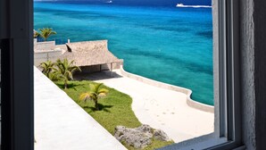 Vista desde una ventana hacia una playa con agua azul cristalina, un barco a lo lejos y una construcción con techo de paja.