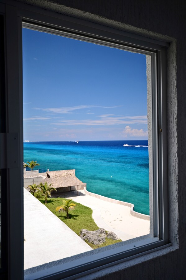 A view from a window overlooking a beach with clear blue water, a boat in the distance, and a thatched-roof structure.