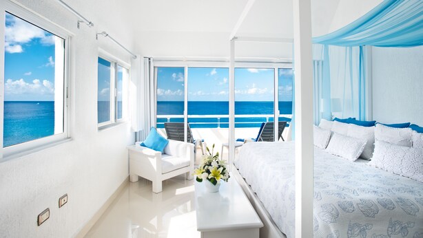 Una habitación luminosa y blanca con un ventanal grande que da al océano, una cama blanca y un sofá blanco con cojines azules.