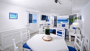Una sala de estar moderna con decoración blanca, una ventana grande con vistas al océano, un comedor con una mesa blanca y sillas azules, y un sofá blanco con cojines azules.