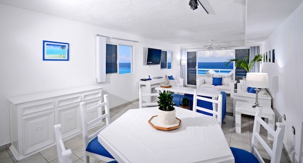 Una sala de estar moderna con decoración blanca, una ventana grande con vistas al océano, un comedor con una mesa blanca y sillas azules, y un sofá blanco con cojines azules.