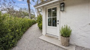 A white building with a gravel path leading to a door, surrounded by greenery and palm trees. - Charming Studio Apartment E - Steps to Beach & Dining (Saint Simons Island)