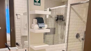 Une salle de bain moderne avec un lavabo blanc, des toilettes, une douche et un espace douche fermé par des parois de verre. Un panneau indique la présence d’une trousse de premiers secours.