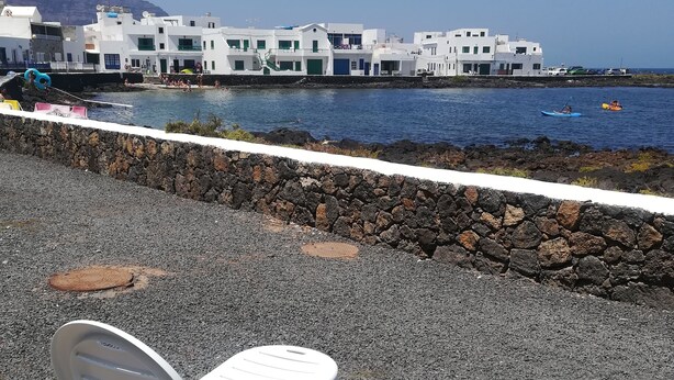 A coastal area with a rocky walkway, two white plastic chairs, and a building with multiple windows in the background. - Brisamarina de Órzola (Orzola)