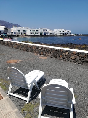 Una zona costera con un paseo marítimo rocoso, dos sillas de plástico blancas y un edificio con múltiples ventanas al fondo.