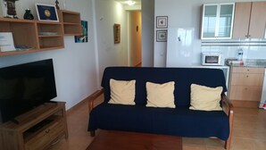 A living room with a dark blue sofa, wooden coffee table, and a TV on a wooden stand. There is a kitchen area with cabinets and a microwave. - Brisamarina de Órzola (Orzola)