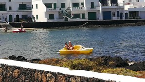 A coastal scene with white buildings, a rocky shoreline, and a yellow boat with two people. - Brisamarina de Órzola (Orzola)