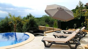 Zona de piscina con sillones de descanso y una sombrilla, rodeada de follaje y vistas a las colinas.