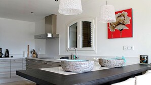 Una cocina moderna con encimera oscura, dos lavamanos de mármol y un amplio ventanal con persianas.
