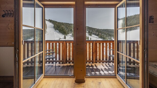Una baita di legno con ampie vetrate che si aprono su un balcone con vista su una montagna innevata.