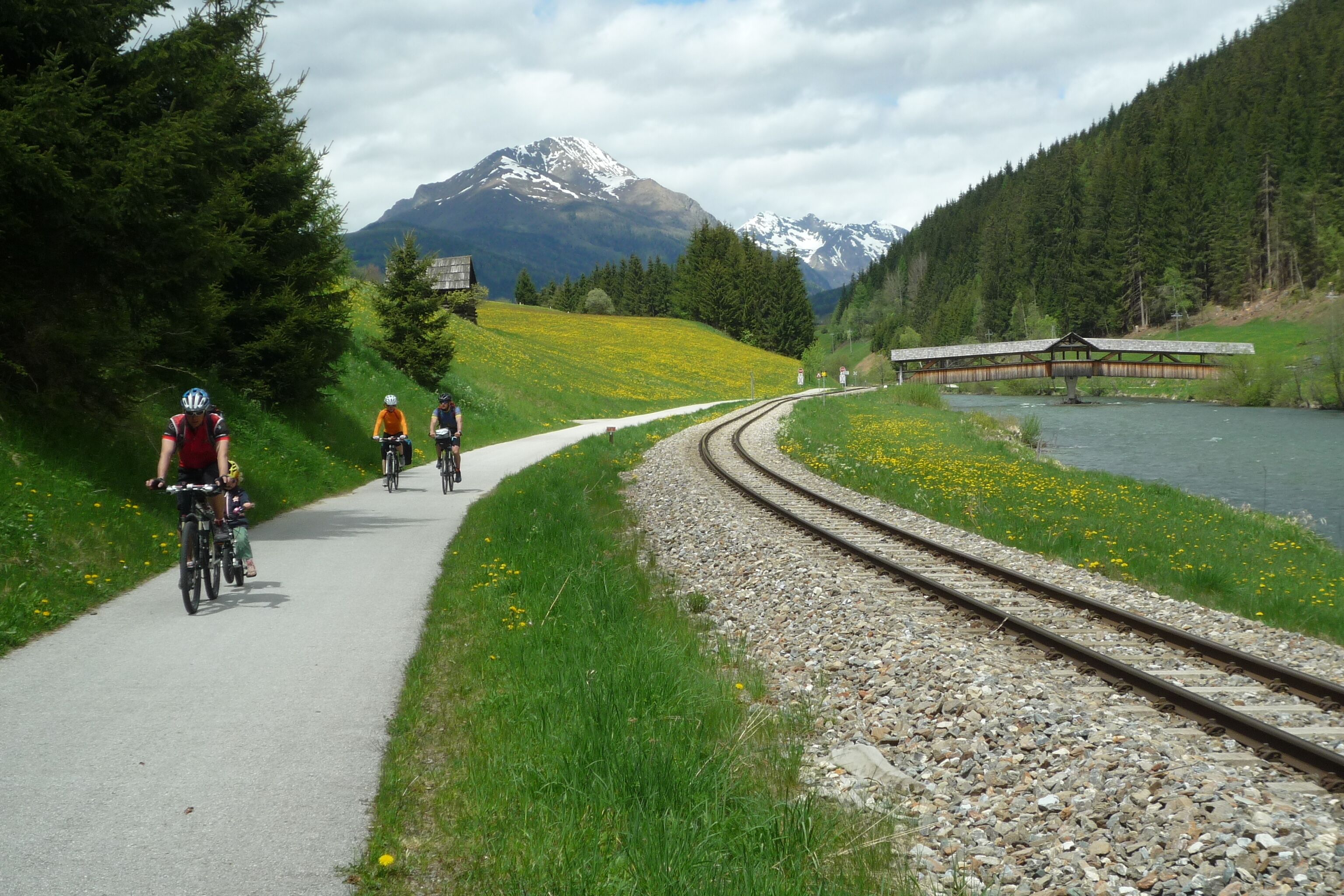 A pathway alongside railway tracks with cyclists, a bridge, and a mountain in the background.