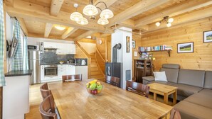 Un accogliente chalet dall'interno in legno, con zona pranzo, un tavolo pieno di frutta, cucina moderna e angolo soggiorno con divano e librerie.
