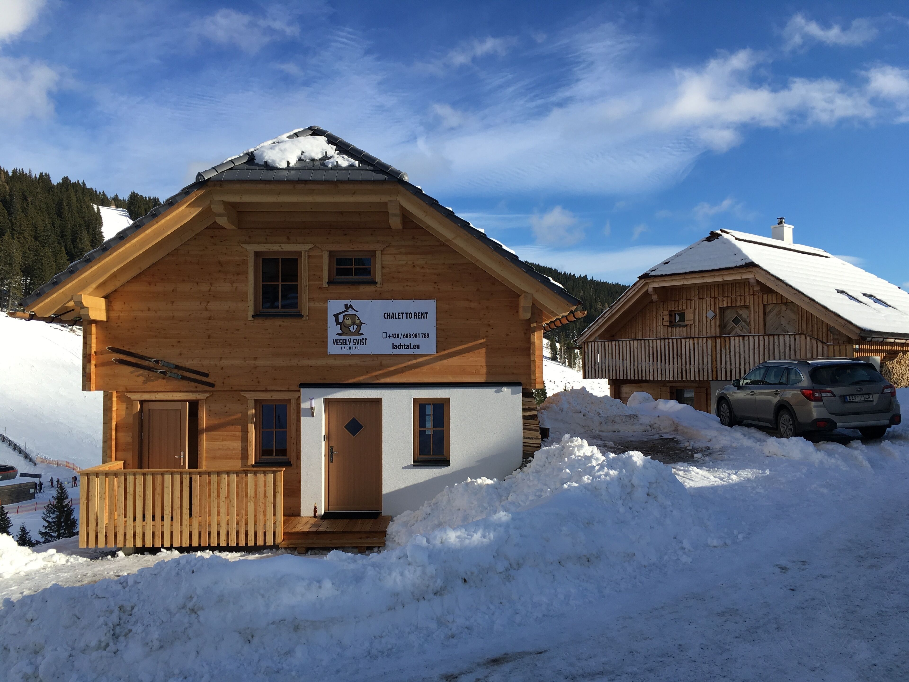 Un chalet de madera con un cartel que indica que está disponible para alquiler, un porche de madera y un coche estacionado cerca.