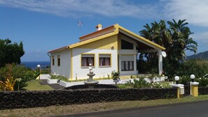A two-story house with a yellow and white exterior, a stone wall in front, and a small garden with a fountain. - HOUSE COAST, on Faial Island, Azores (HORTA)