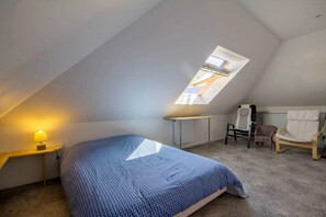A bedroom with a sloped ceiling, a skylight, a bed with a patterned bedspread, a bedside lamp, a chair, and a radiator.
