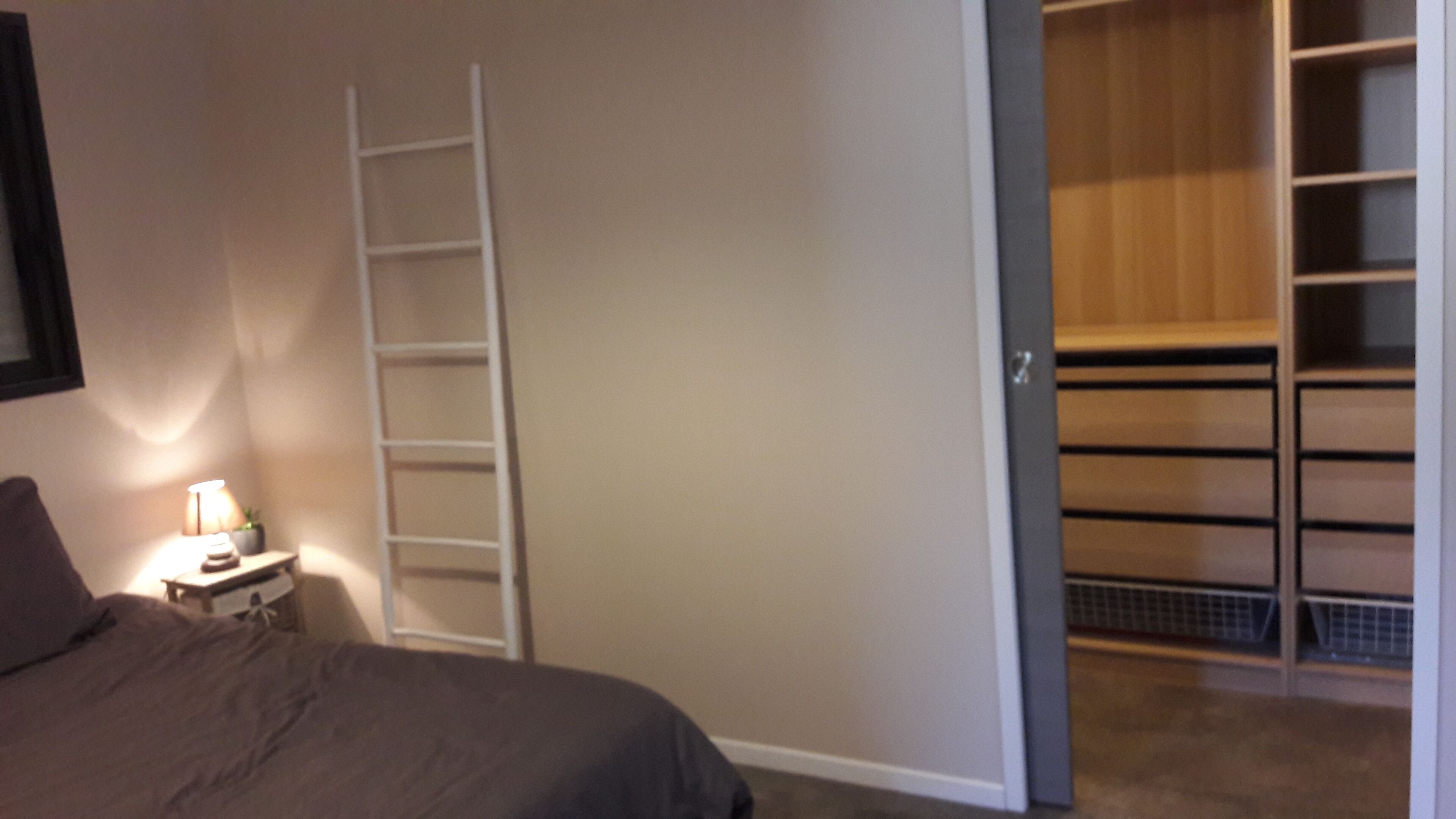 A bedroom with a bed, a nightstand with a lamp, a white ladder, and a closet with shelves.