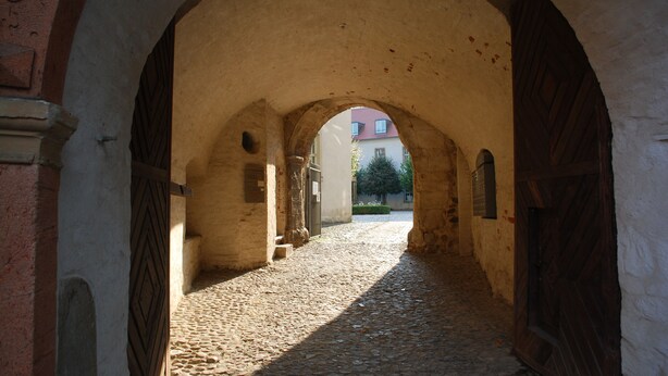 Un couloir en arc, dont le sol est pavé de pierres, mène à un bâtiment surmonté d’un toit rouge.