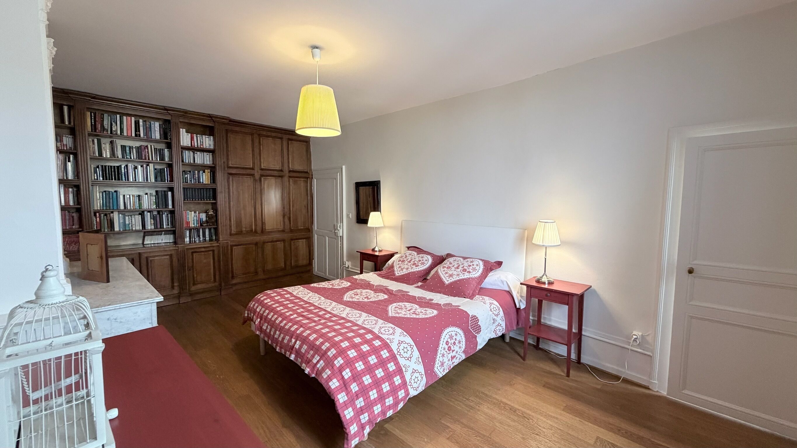 A bedroom with a large bed, two bedside tables with lamps, a wooden bookshelf, and a white door.