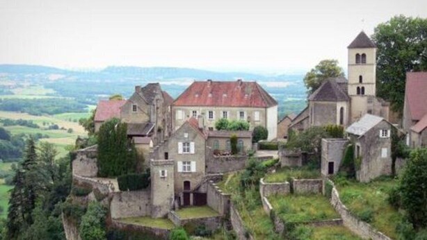 Edifici de pedra històric amb diverses estructures, incloent-hi una torre, envoltat de vegetació exuberant i un paisatge amb turons.
