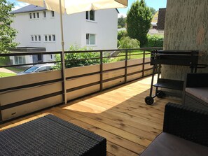 Um deck de madeira com móveis de exterior, guarda-corpo de metal e vista para um edifício branco ao fundo.