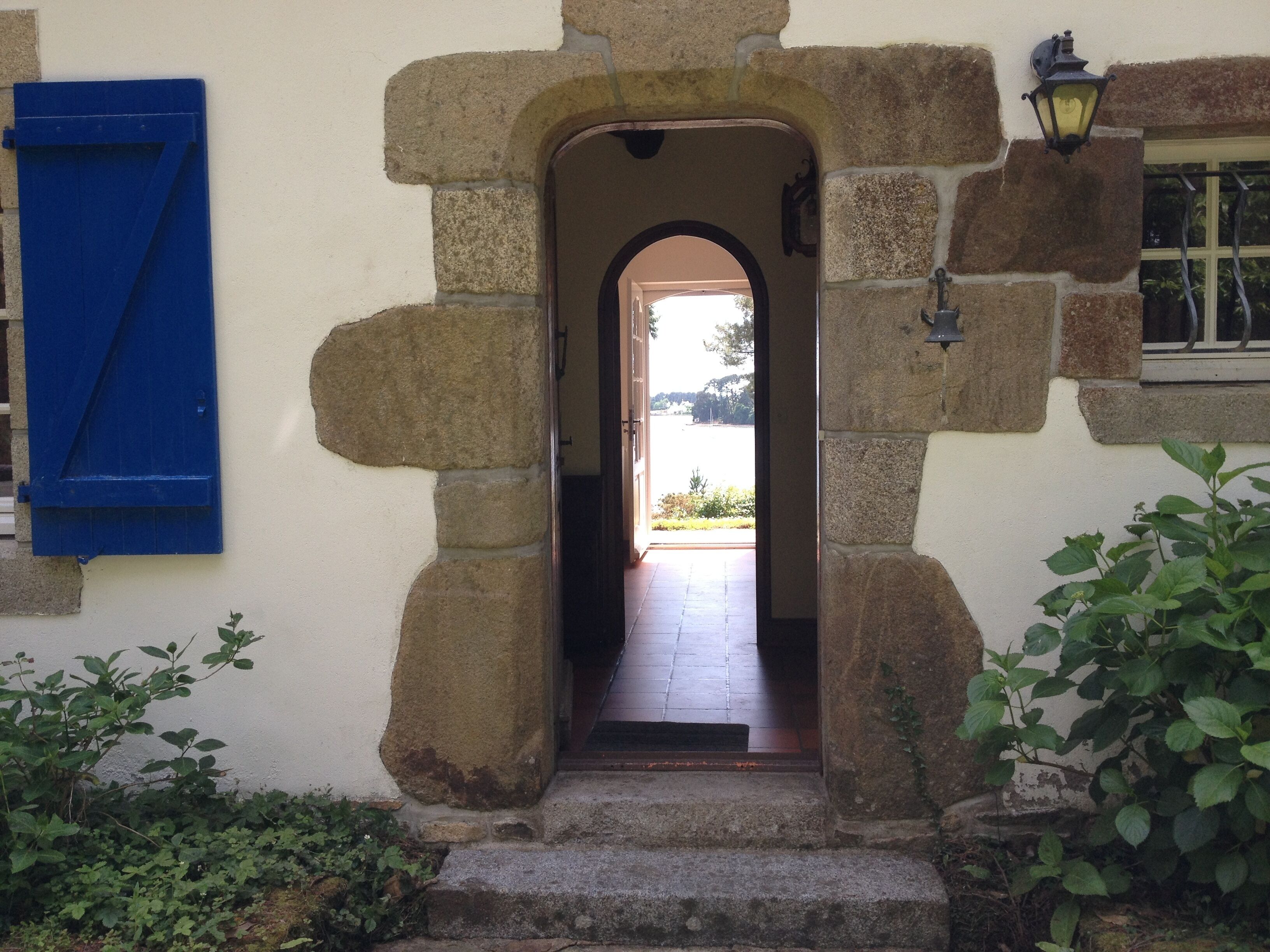 A stone entrance with a blue shutter, a lantern, and a view into a room with a tiled floor.