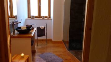 Een compacte badkamer met een houten vloer, een klein wastafelmeubel, een handdoekhouder, een raam met daglicht en een douchehoek.