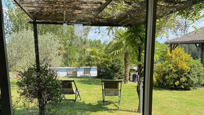 Une terrasse couverte avec vue sur une piscine, une table en bois agrémentée d’un vase décoratif, et une pelouse verdoyante avec un petit arbre et un banc.