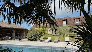 Un espace piscine avec des chaises longues, une terrasse en bois et, au fond, un bâtiment doté d’un toit recouvert de tuiles.