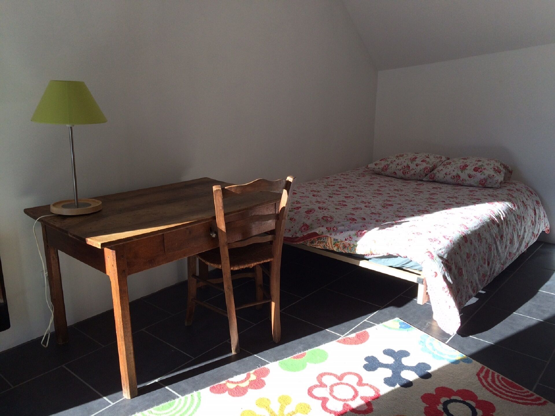 A bedroom with a wooden desk, a green lamp, a bed with floral bedding, and a colorful rug.