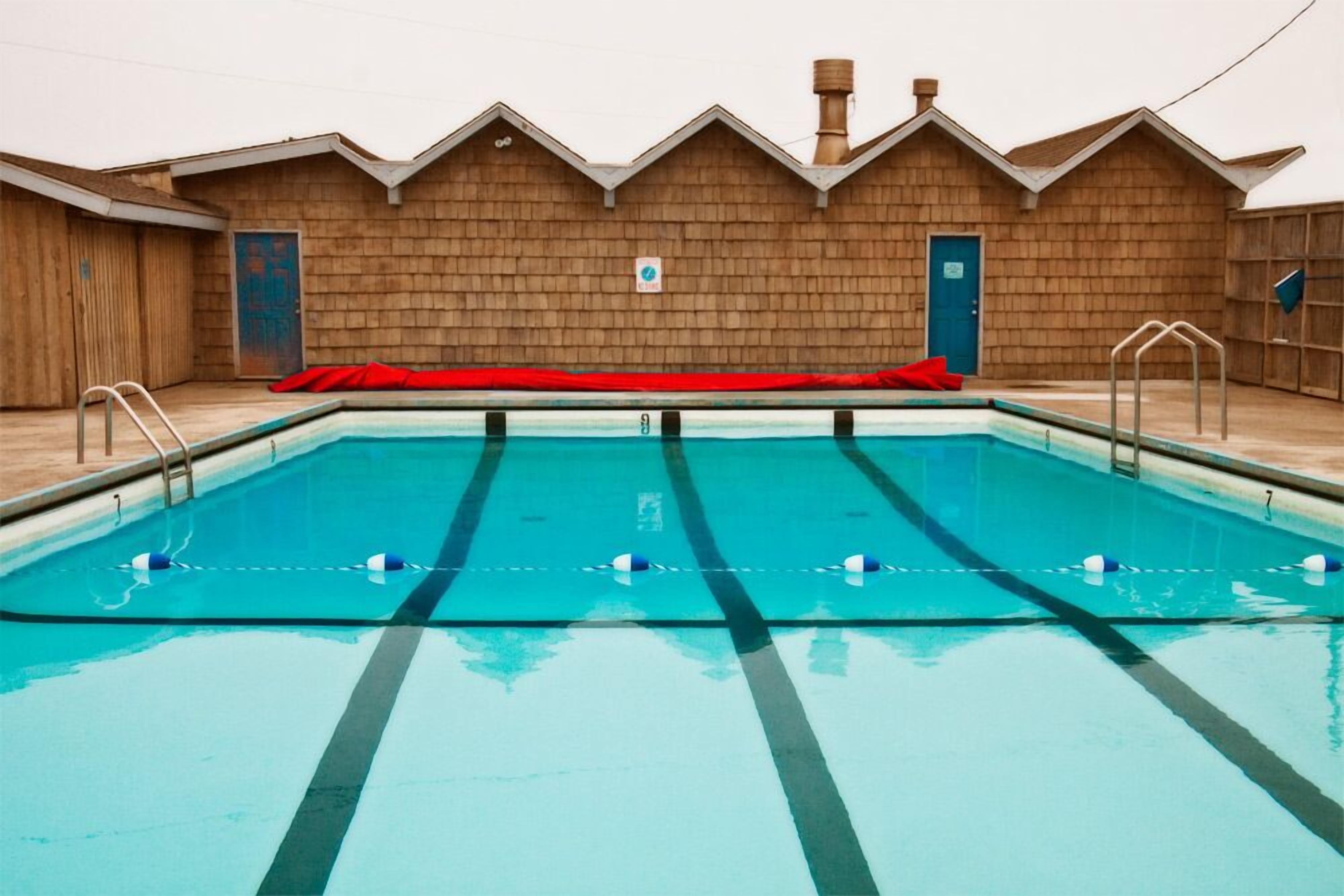 A rectangular swimming pool with lane dividers, a red deck, and a wooden building with a peaked roof in the background.