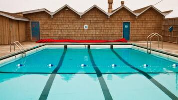 A rectangular swimming pool with lane dividers, a red deck, and a wooden building with a peaked roof in the background.