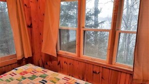 Ein Zimmer mit einer Holzwand, einem Bett mit farbenfroher Steppdecke und einem Fenster mit Blick auf Bäume.