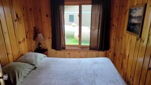 A wooden room with a bed, a window with curtains, a lamp, and a painting on the wall. - Bayview Fireside Cabin (Tower)