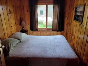 Una habitación de madera con una cama, una ventana con cortinas, una lámpara y un cuadro en la pared.