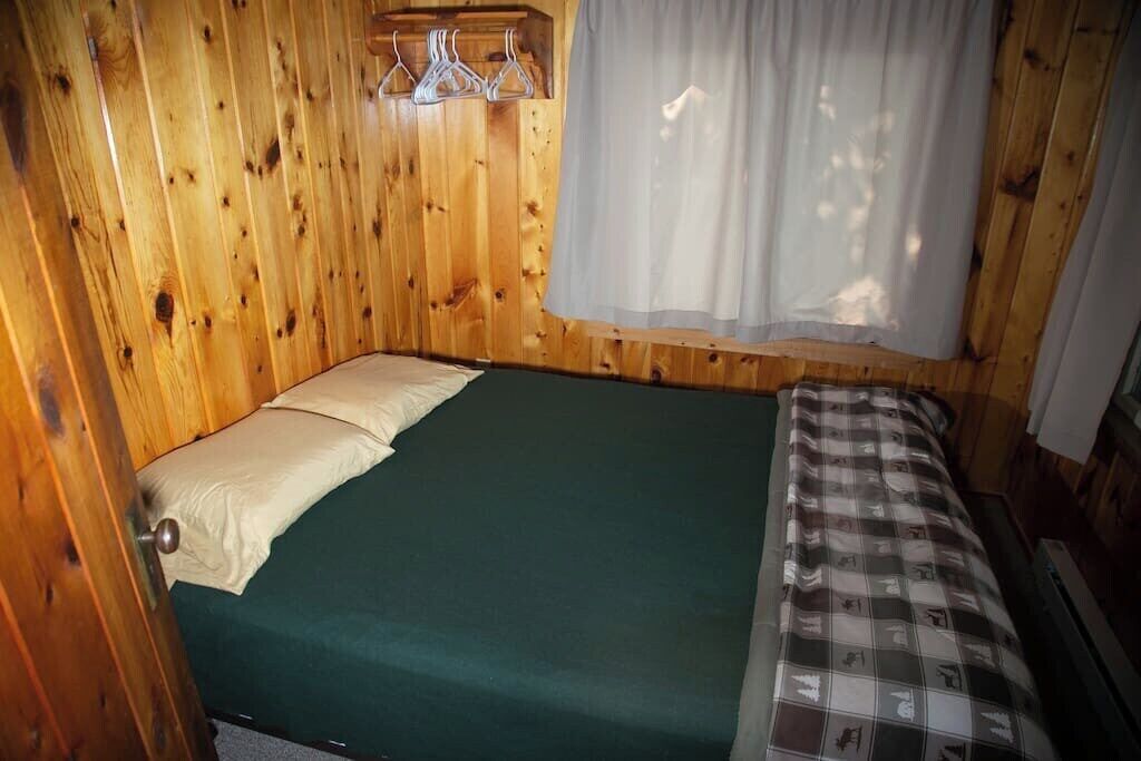 A room with wooden walls, a bed with a green mattress, and a window with a white curtain.