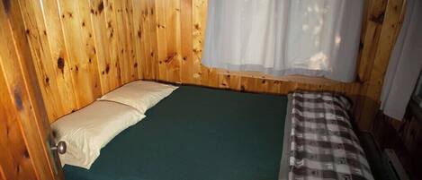 A room with wooden walls, a bed with a green mattress, and a window with a white curtain.