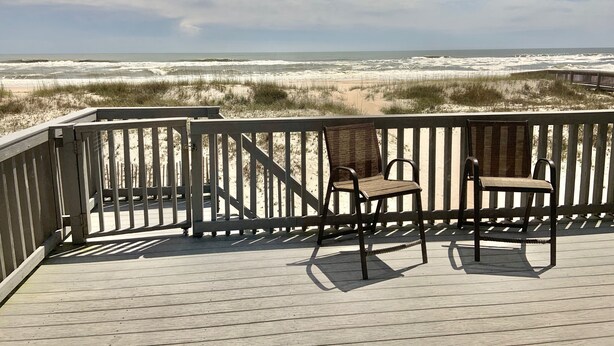 Eine Holzterrasse mit zwei Korbstühlen mit Blick auf einen Strand mit Dünen und Wellen.