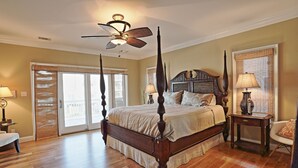 A spacious bedroom with a four-poster bed, a ceiling fan, and a large window with blinds. - The Ultimate In Beach Living! Ocean View, Steps to Beach,  Sleeps 8. Elevator! (Tybee Island)