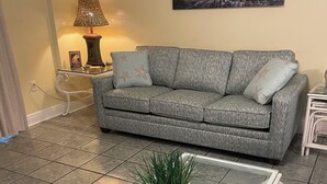A living room with a grey sofa, a glass coffee table with a plant, and a painting of a coastal scene. - AUGUST REDUCED/SEPTEMBER AVAILABLE!! (Orange Beach)
