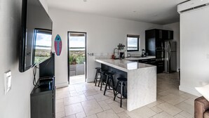 A modern kitchen with a large island, bar stools, a flat-screen TV, and a view of the beach.
