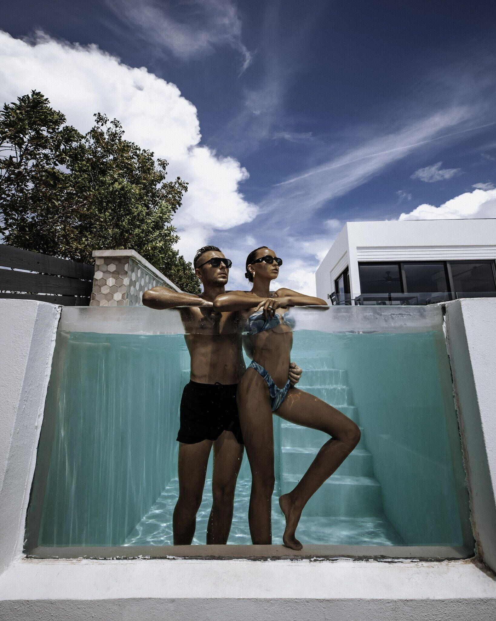 En mand og en kvinde i badetøj står i en swimmingpool med en moderne bygning i baggrunden.