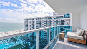 Un balcón moderno de hotel con piscina, zona de estar y vistas al océano y otros edificios.