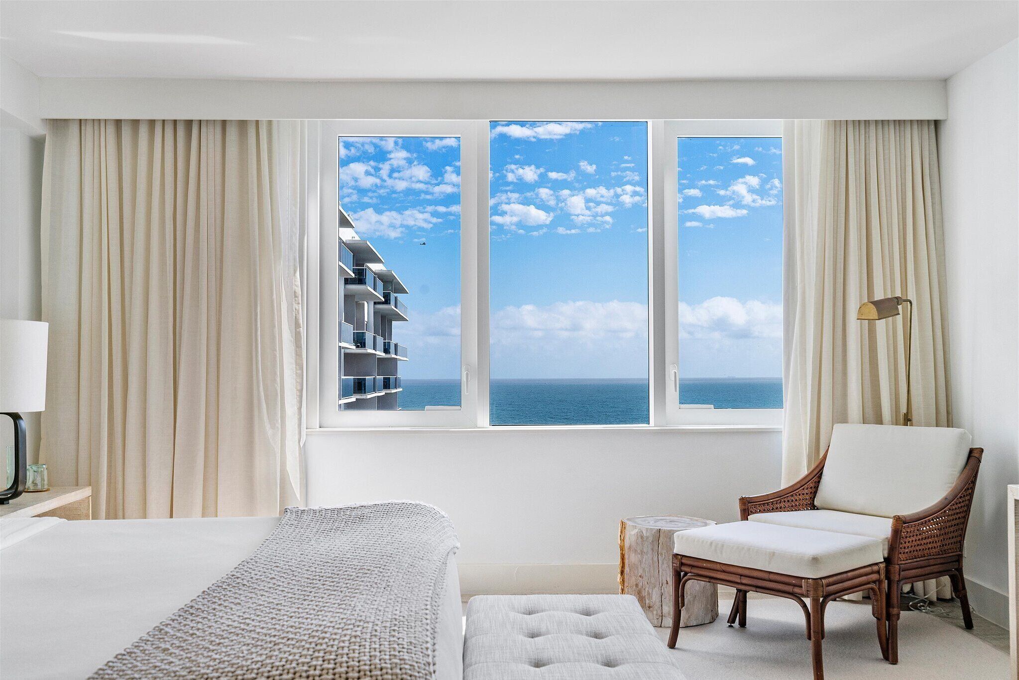 A hotel room with a large window offering a view of the sea and a building. There is a bed, a chair, and a small table.
