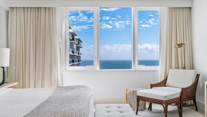 Habitación de hotel con una ventana grande que ofrece vista al mar y un edificio. Hay una cama, una silla y una mesita.