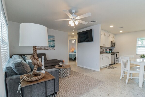 A modern living room with a grey sofa, a coffee table, and a ceiling fan. A kitchen with white cabinets and a dining area with a white table and chairs is visible in the background.