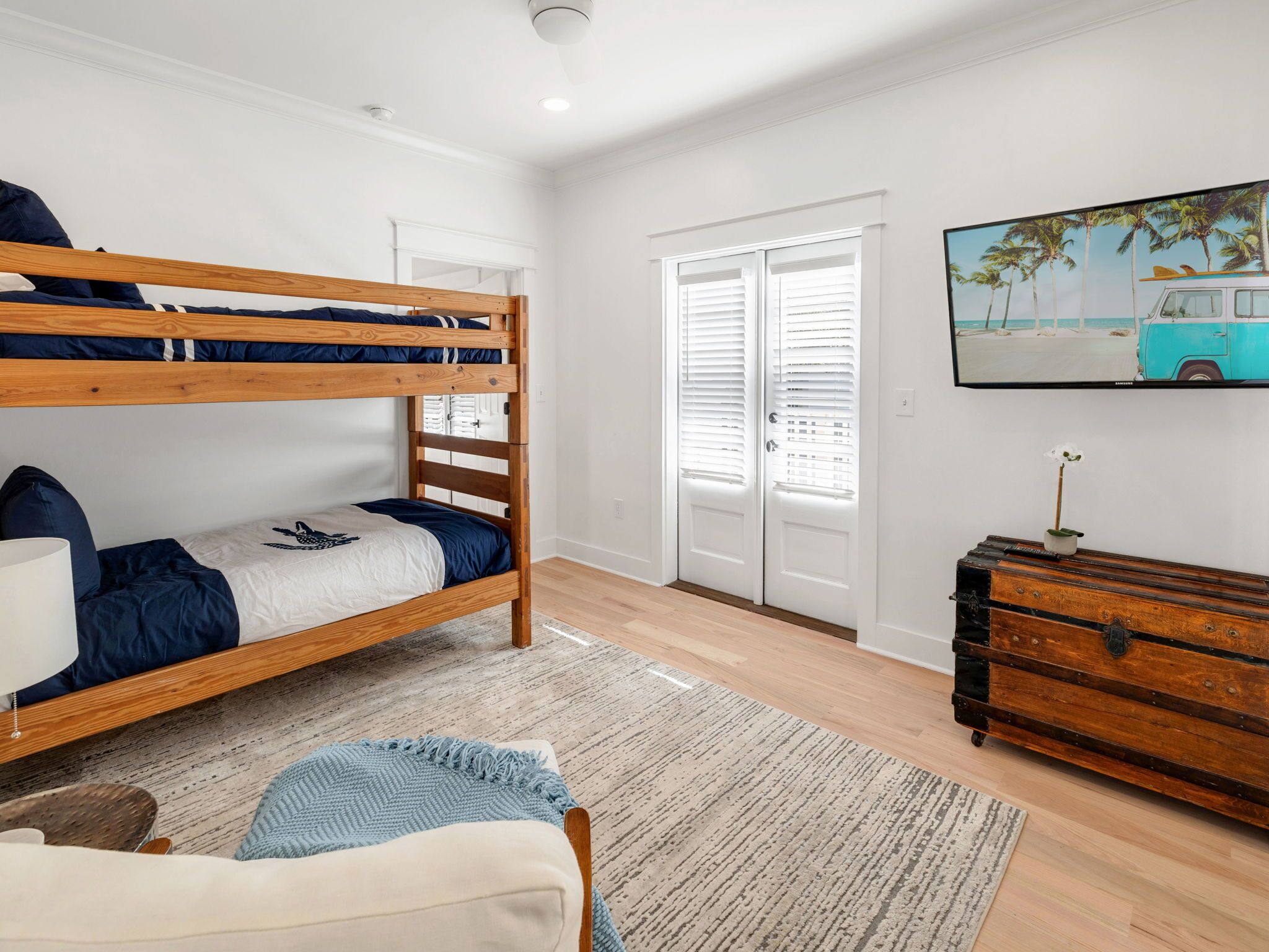 Uma cama beliche com estrutura de madeira, uma pequena mesa de cabeceira com luminária, um tapete azul e branco estampado, um aparador de madeira com uma planta e um quadro de um carro azul na parede.