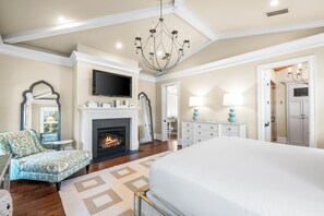 A spacious bedroom with a large bed, a fireplace, a television, and a chandelier. - Elegant Camp District Home with Electric Cart, 6 Bikes & Watercolor Beach Club Access (Santa Rosa Beach)