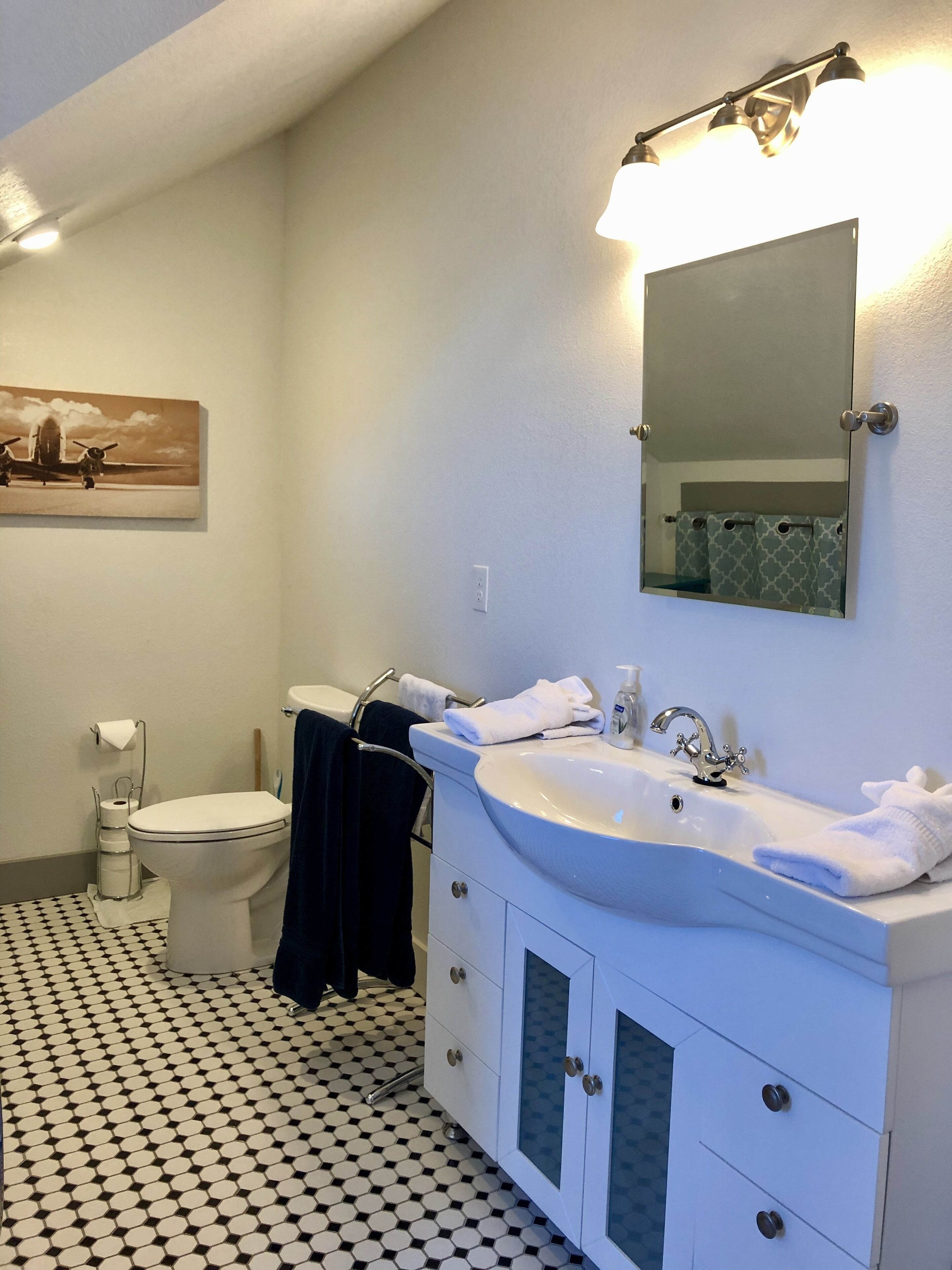 Un baño con un lavamanos blanco, espejo y inodoro. El piso presenta un diseño blanco y negro y hay un cuadro en la pared.