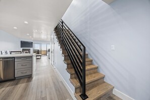 A modern kitchen with a stainless steel dishwasher, a wooden staircase with black railings, and a view to a dining area.
