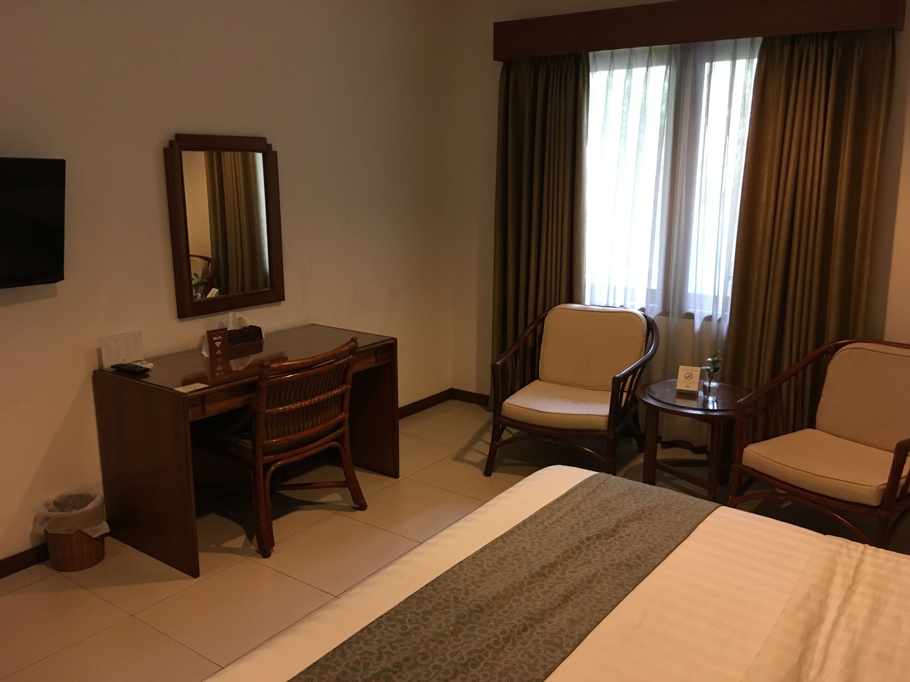 A hotel room with a bed, a desk, two chairs, a mirror, and a window with curtains.